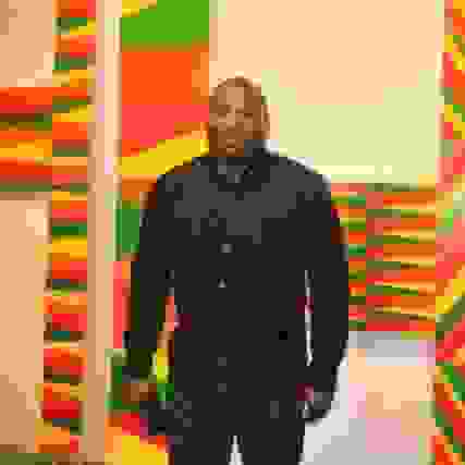 Rico Gatson in a black dress shirt standing in front of a colorfully patterned artwork