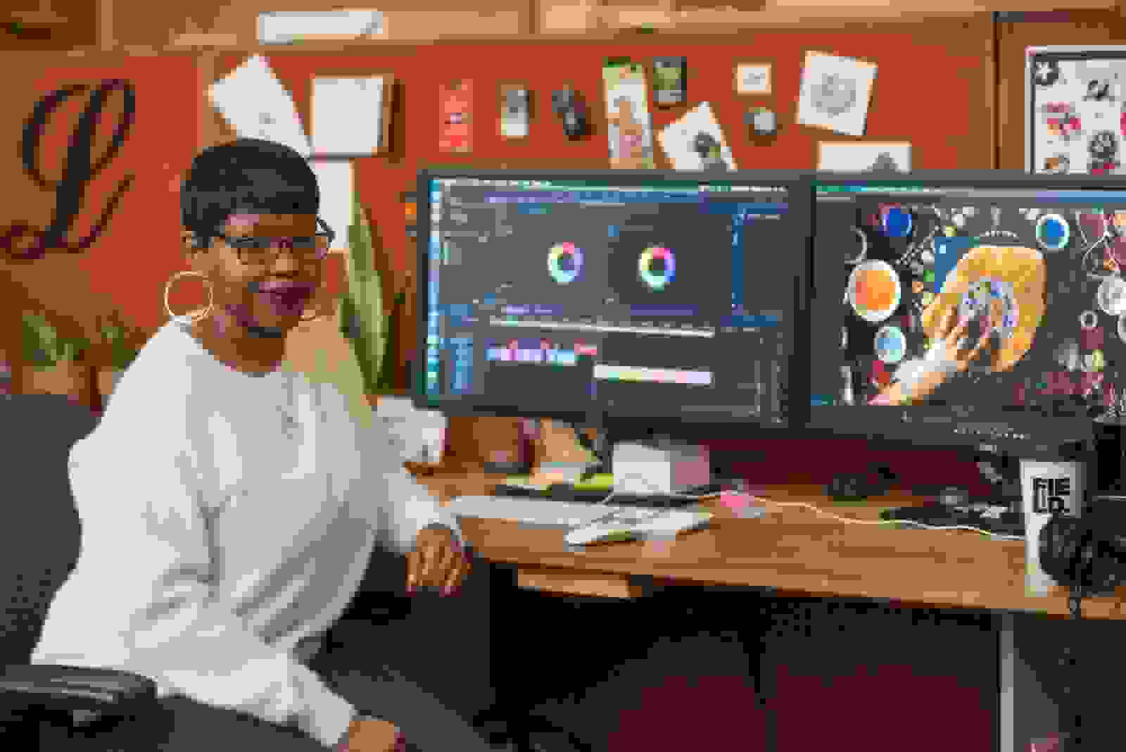 Latoya Flowers sitting at her desk, with two monitors displaying the Adobe Premiere program workspace.
