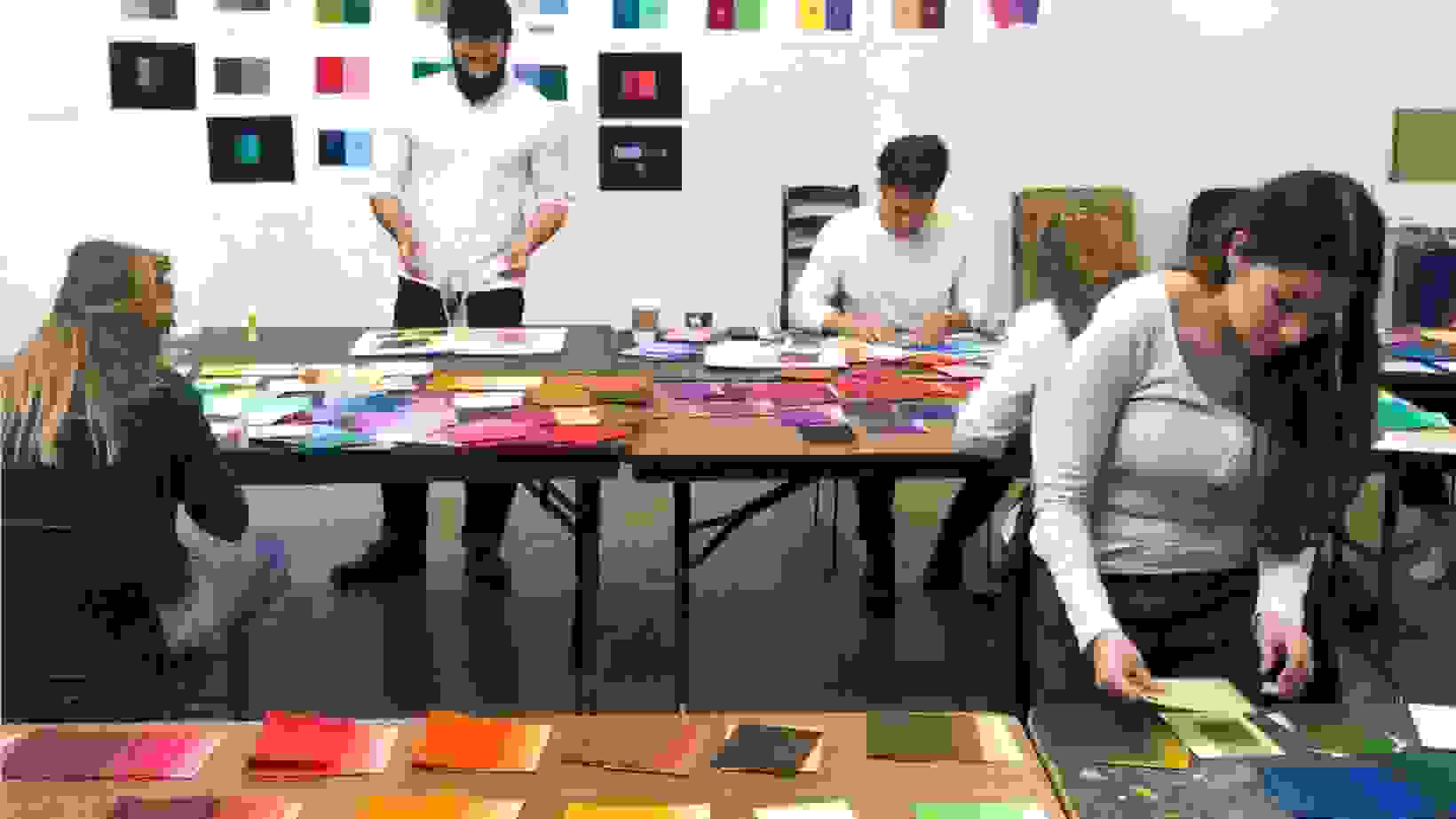 Students in a classroom work on color theory exercises, using sheets of colored paper.