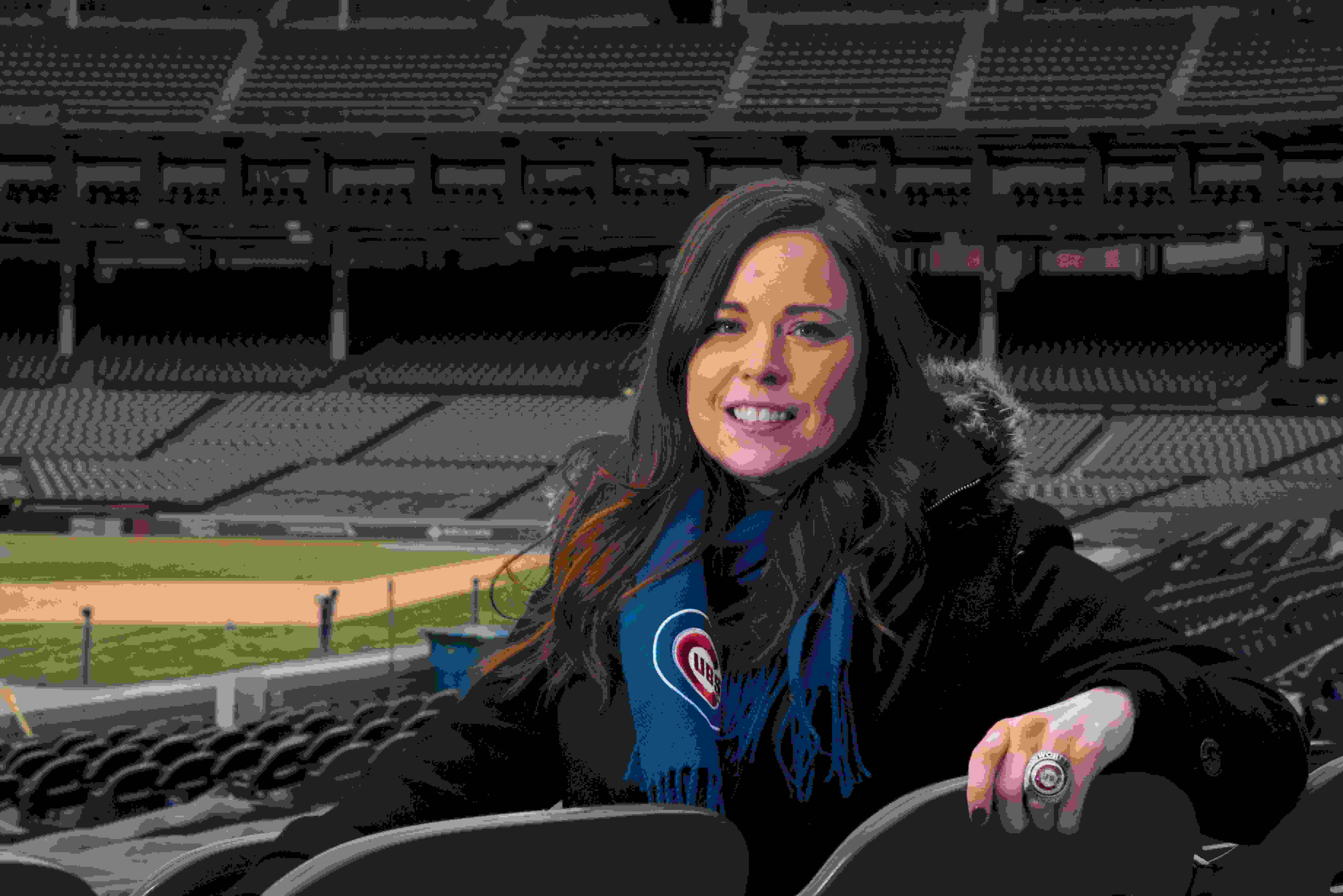 Kelly King sits in a green plastic seat in Wrigley Field. You can see the empty stadium behind her. She is smiling wearing a blue scarf with the Cubs logo and a large World Series ring on her left hand