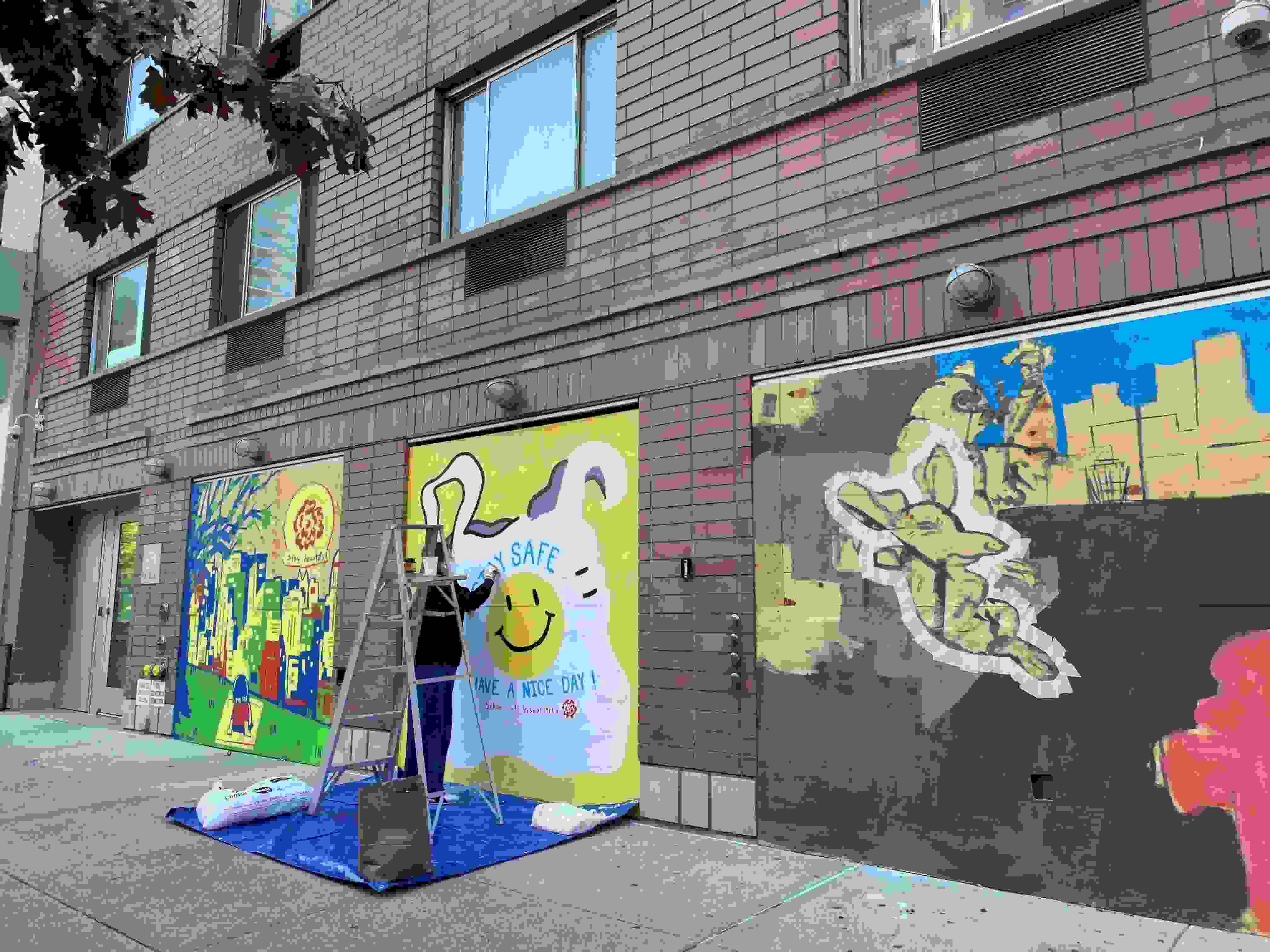 A photograph of a city building exterior, on which are three murals painted on plywood at the street level. A person is working on the middle mural.