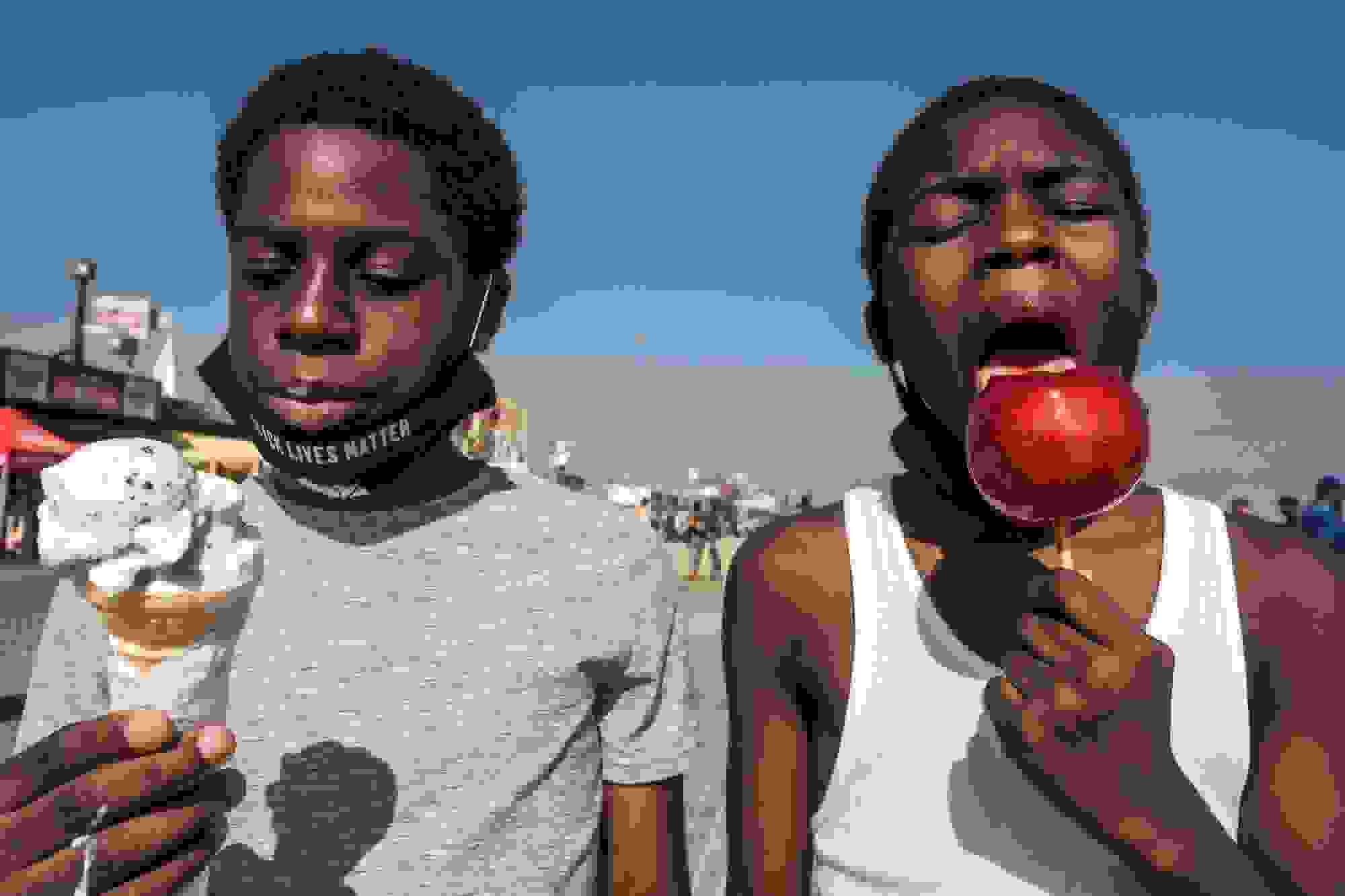 A street photograph on a boardwalk on a sunny day. Taking up the foreground are two young Black men side by side. The one on the left is looking down at the ice cream cone he is holding. The one on the right is in mid-bite of a red candy apple.
