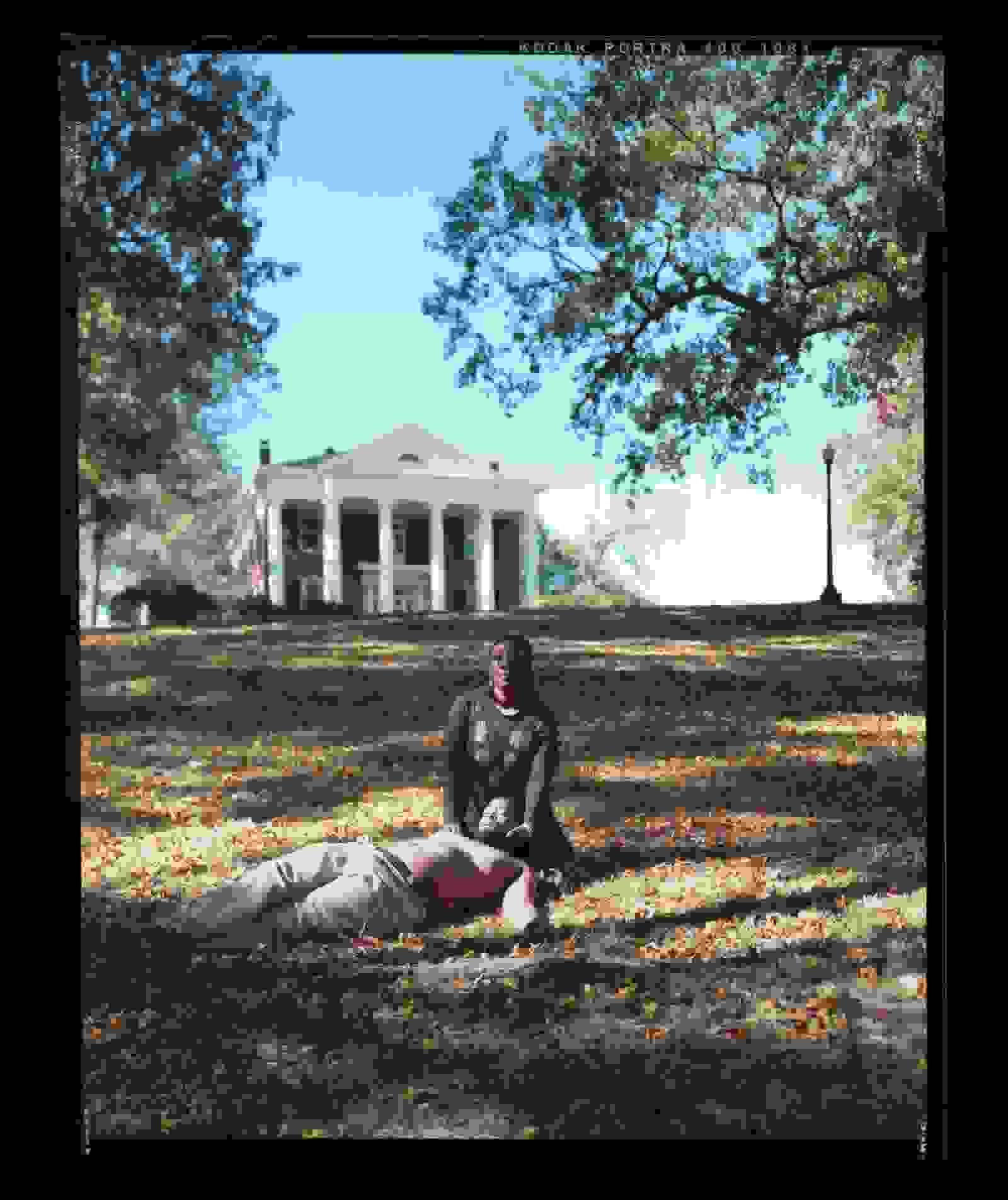 A color photograph of a black woman sitting in the grass cradling the head of a shirtless black man on her lap, as he lies flat in the grass. In the background is a brick building with white columns, trees, blue skies, and a streetlamp.