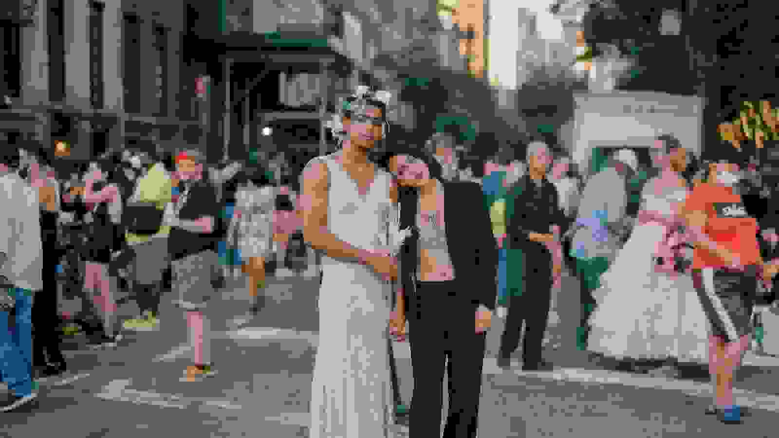 Two people are holding each other in the middle of a crowded street. One person is wearing a dress with a flower crown and the other a suit. 