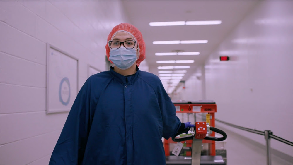 A still of a person in scrubs and a mask walking down a hallway while wheeling a pallet of hospital materials.