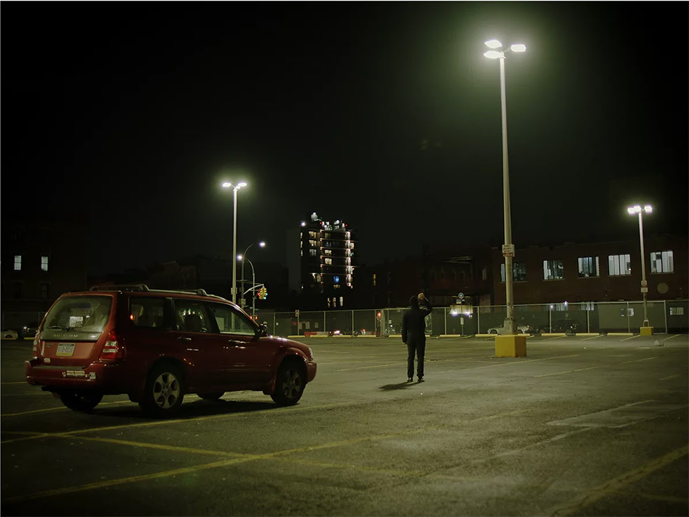 A man standing outside of a red car in a parking lot at night. 