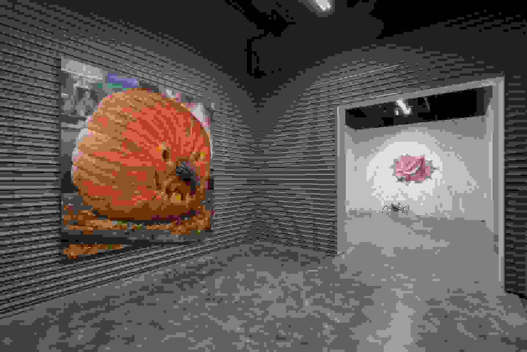 A grey room with a painting of a carved pumpkin on the wall, and another room in the distance with a painting of a woman's mouth pointing down towards a halloween decoration spider, surrounded by webs.